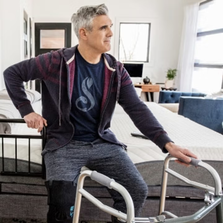 A man sitting on the edge of a bed with a walker nearby